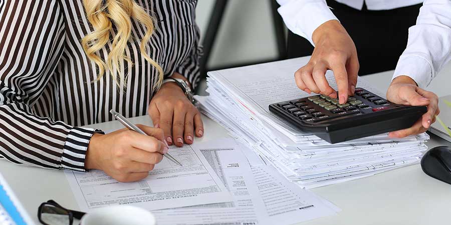 People using a calculator  with a stack of paperwork on a desk.